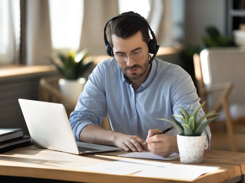 Focused young businessman in eyeglasses wearing wireless headset with microphone, involved in online video call negotiations meeting with partners colleagues or studying distantly, writing notes.