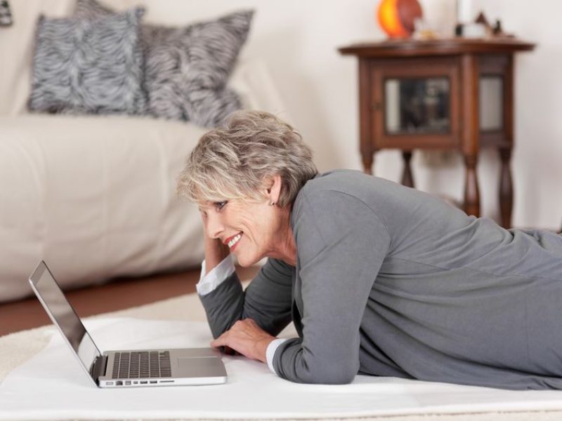 21190330 - image of an elderly happy woman lying on the floor and working on the laptop.